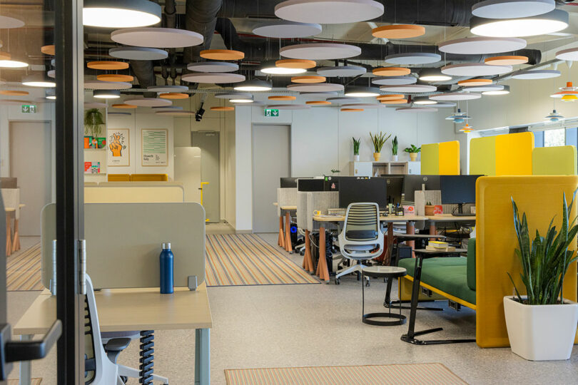 Modern open-plan office with desks, chairs, computers, and acoustic ceiling panels. Potted plants and colorful partitions add decor. Bright lighting and natural sunlight illuminate the space.