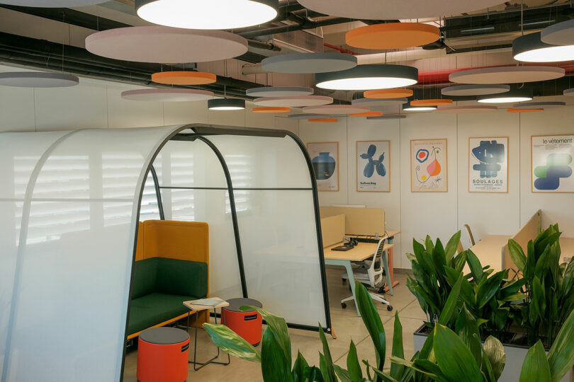 Modern office with colorful ceiling panels, white partition, seating area, desks, potted plants, and wall art.