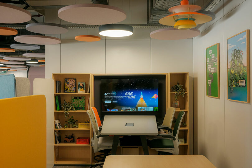 Modern meeting room with a central table surrounded by chairs, a wall-mounted screen displaying a presentation, and shelves with books and plants. Circular ceiling panels add color.