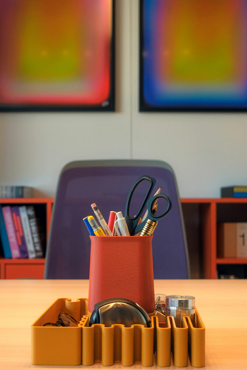 A desk with an organizer holding scissors, pens, and pencils. Blurred artwork hangs on the wall. Books are visible on shelves in the background.