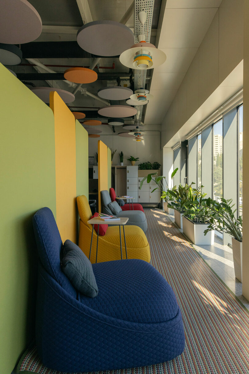 A modern seating area with colorful chairs and dividers, ceiling decorations, and large windows allowing natural light. Potted plants line the windowsill.