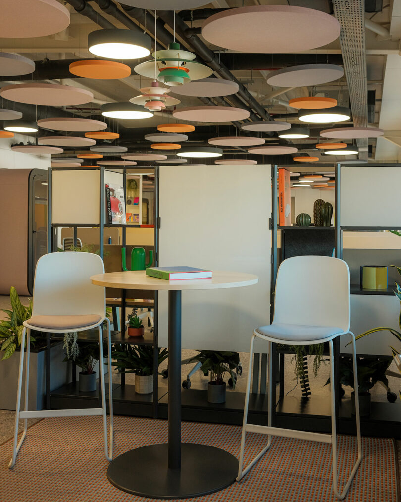 Modern office space with two white chairs at a round table. Background features partition with decorative plants and circular ceiling lights.