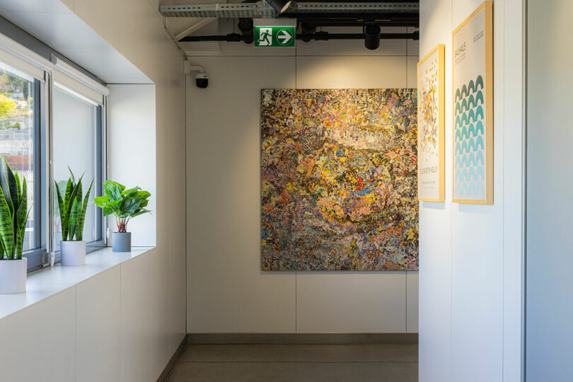 Hallway with a large abstract painting on the wall, two framed pictures, potted plants on a windowsill, and a ceiling-mounted exit sign.