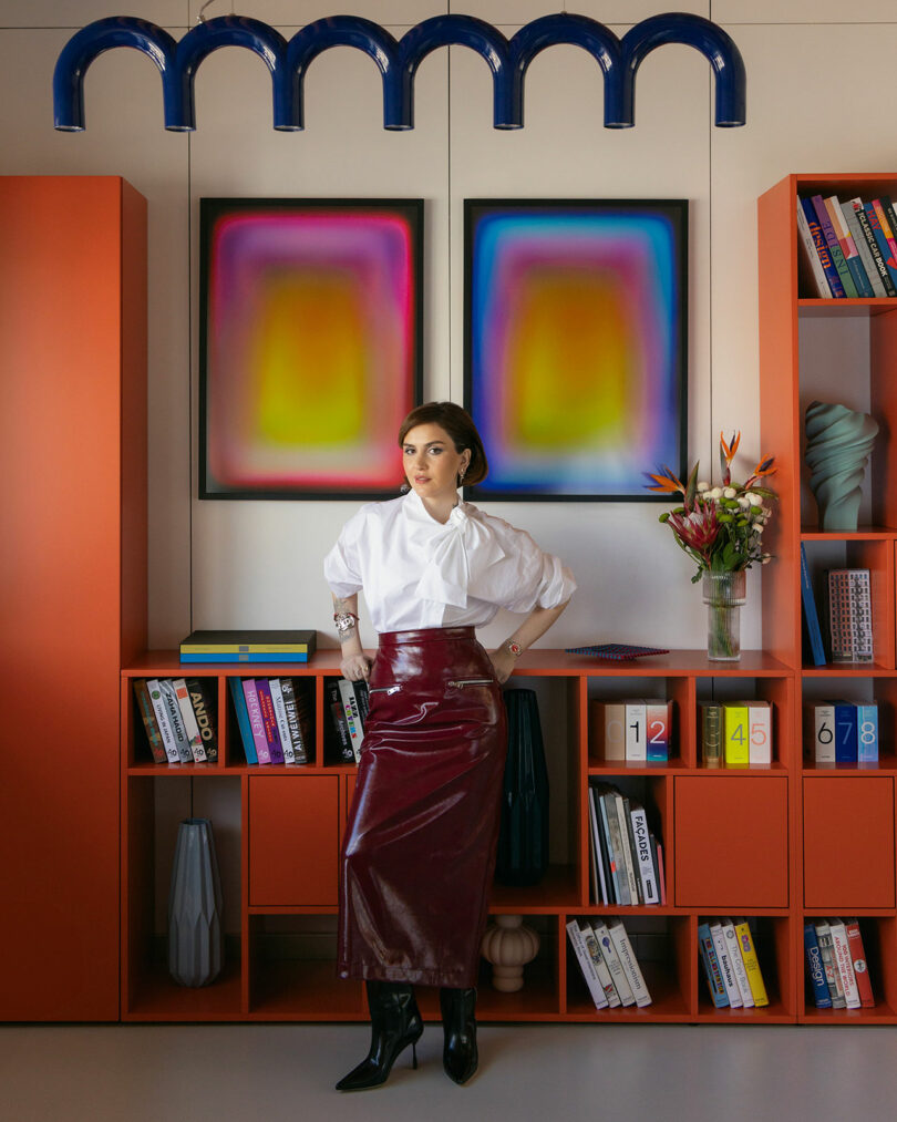 Woman in a white blouse and red vinyl skirt stands in a modern room with red shelving, colorful artwork, and books.