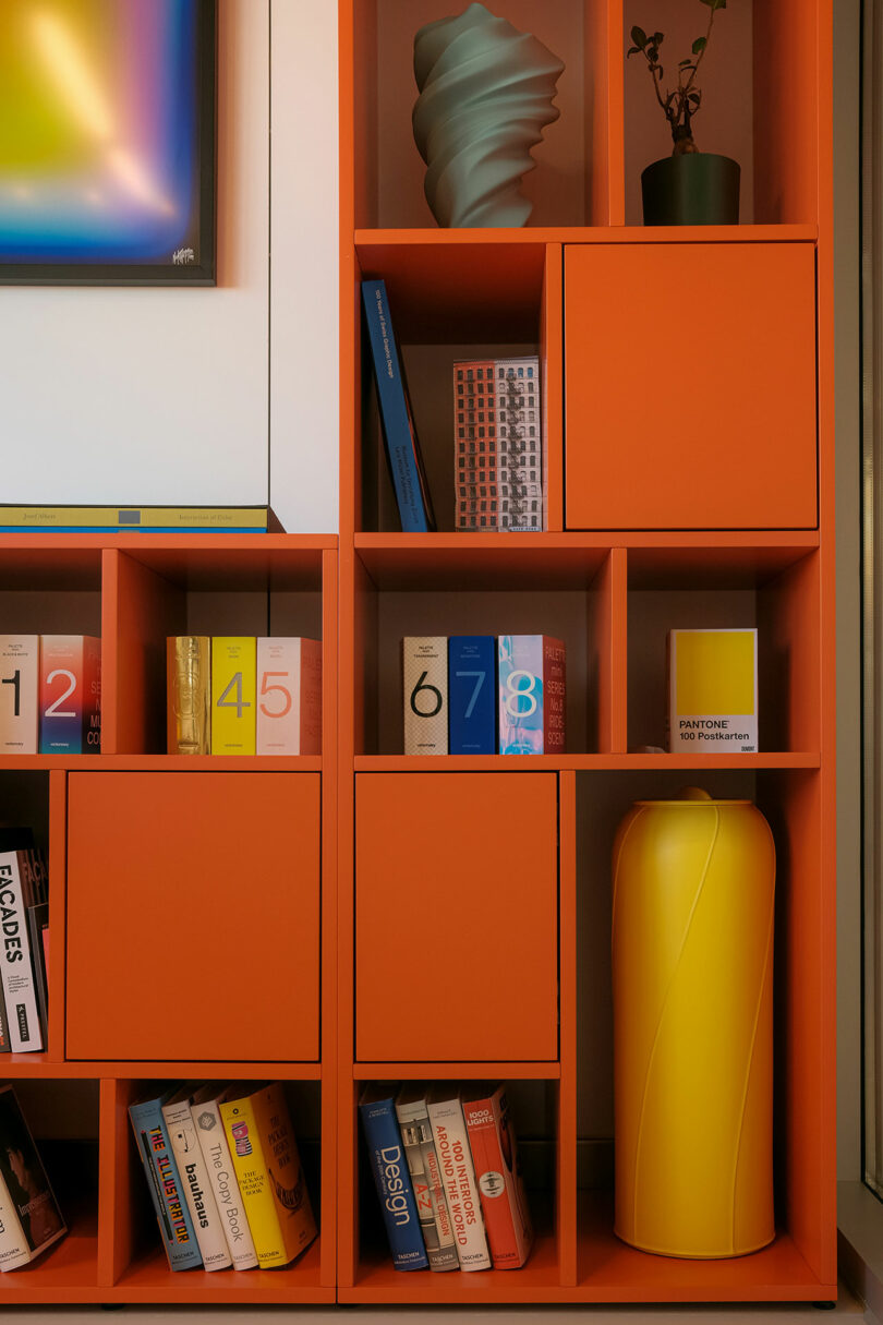 Orange shelving unit with books, colorful boxes, a yellow vase, a green abstract sculpture, and a potted plant. A corner of framed art is visible on the top left.