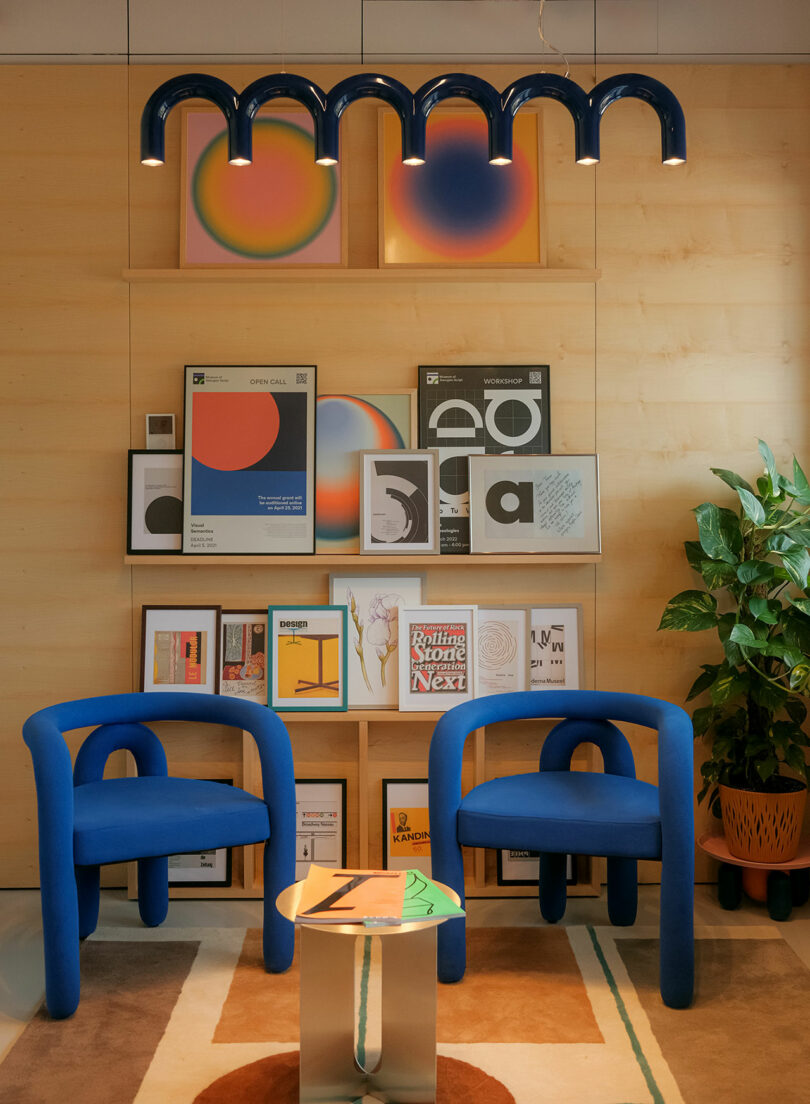 Gallery with two blue chairs, a low table, and artwork on the wall. A blue, arched light fixture hangs above. There is a potted plant on the right.