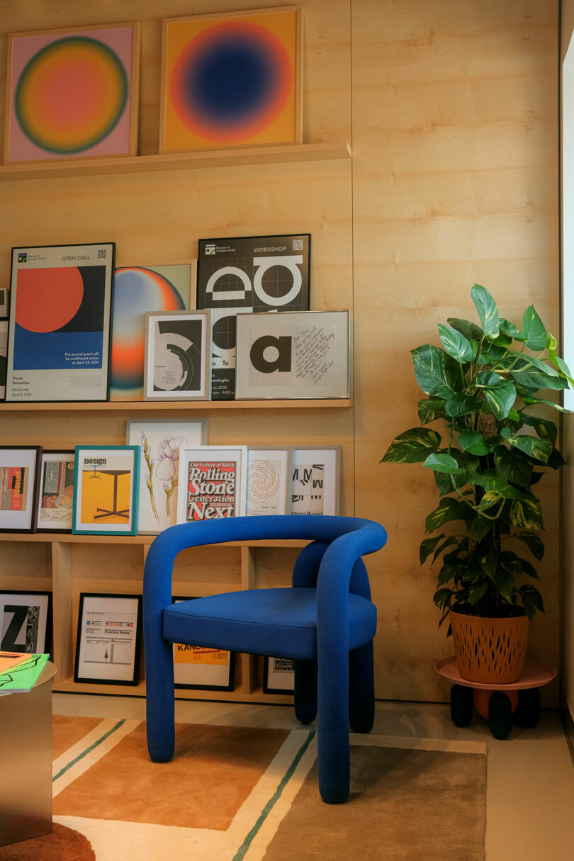 A room with a blue chair, a tall plant, and artwork on wooden walls. The art includes abstract designs and typography. A window allows natural light into the space.