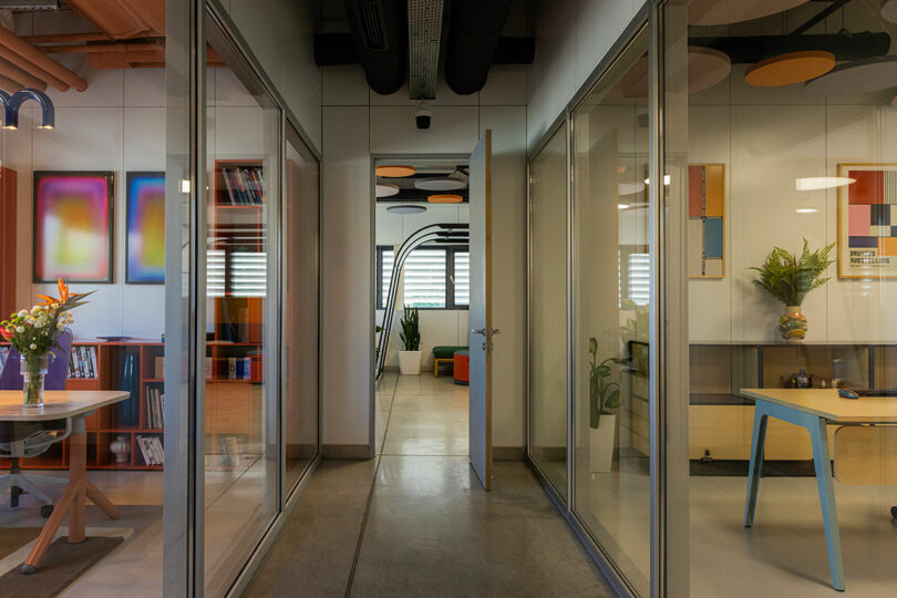 Modern office hallway with glass walls, open doors, and framed art. Plants decorate the space, which includes desks and shelves. Bright lighting.