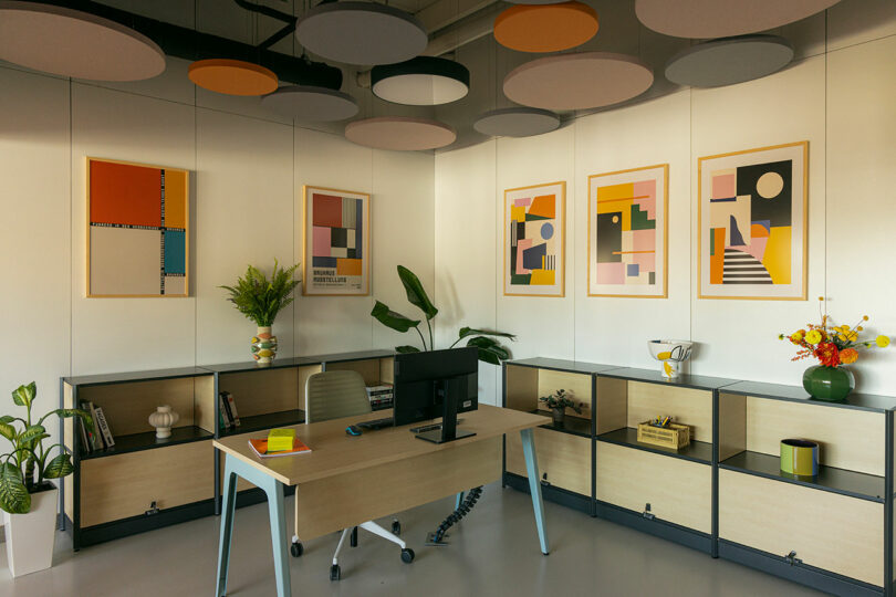 Modern office with a wooden desk, computer, and chair. Geometric art on walls, plants, and shelves with books and decor items. Circular ceiling panels in various colors.