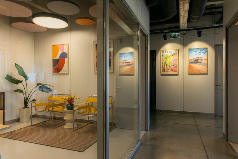 Modern office hallway with framed art on the walls and a glass-walled seating area. Inside, there are yellow chairs, a white table, and a potted plant on a beige rug.