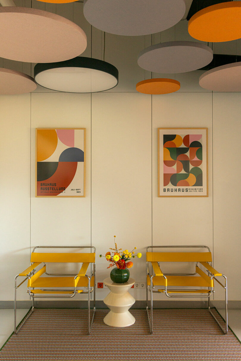 Two yellow chairs and a vase with flowers on a small table are arranged against a wall with geometric art. The ceiling features hanging circular panels in various colors.