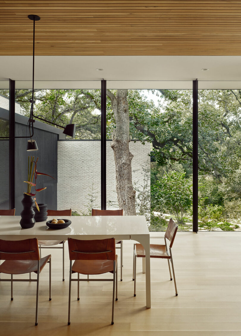 Comedor moderno con una mesa blanca rectangular y cinco sillas marrones. Un jarrón negro con ramas se encuentra en la mesa. Las ventanas grandes revelan una vista de los árboles y una pared de ladrillo blanco afuera.