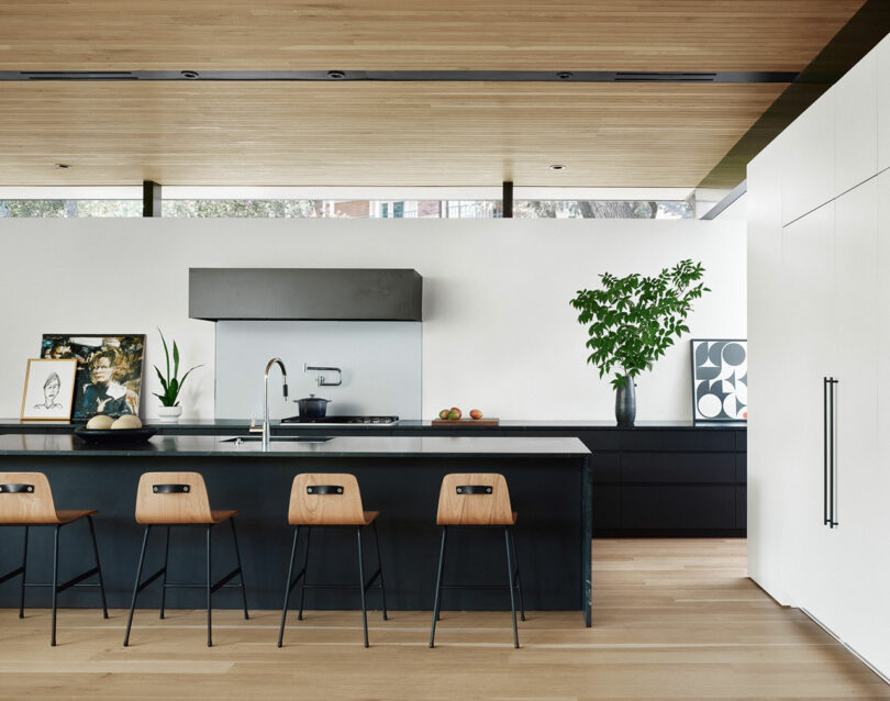 Modern kitchen with a long black island, four wooden bar stools, artwork, a potted plant, and a sleek design. Light wooden ceiling and floor add warmth to the minimalist setting.