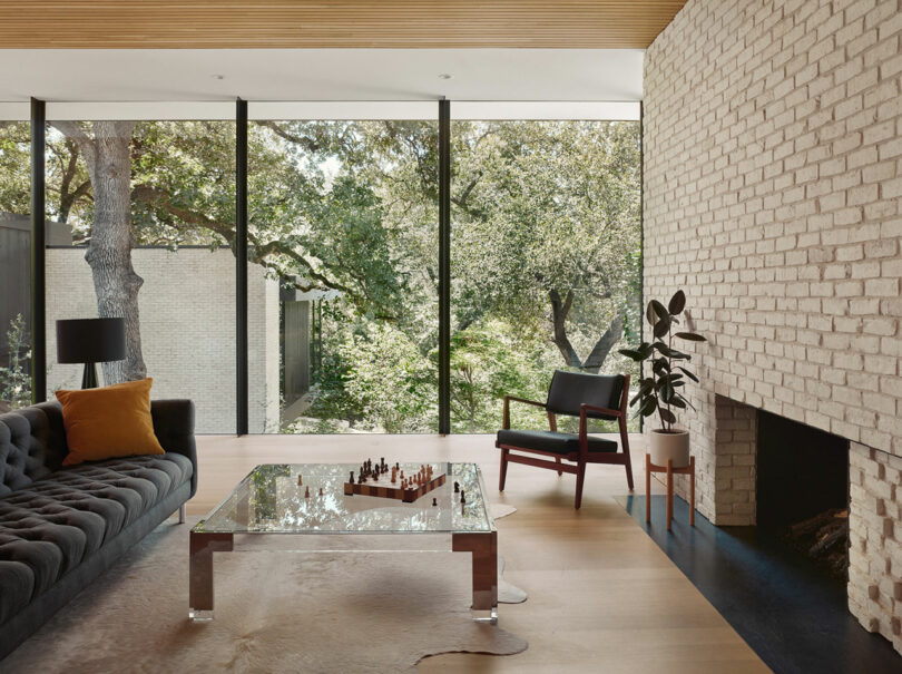 Sala de estar moderna con paredes de ladrillo, grandes ventanas que muestran vegetación, una mesa de café de vidrio con un ajedrez, un sofá oscuro con una almohada naranja, una silla negra y una planta junto a la chimenea.