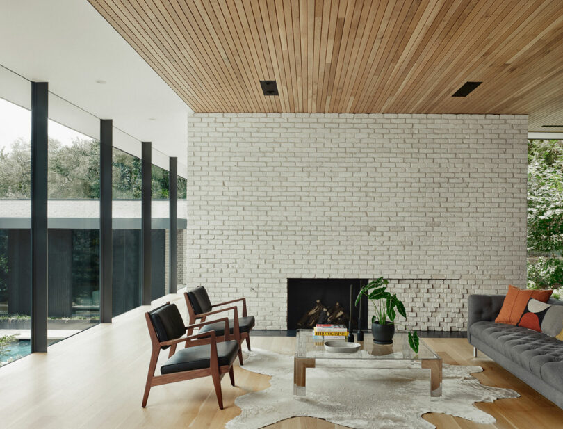 Sala de estar moderna con una pared de ladrillo blanco, chimenea, sofá gris, sillas de madera y alfombra de piel de vaca. Las ventanas grandes revelan una vista de los árboles.