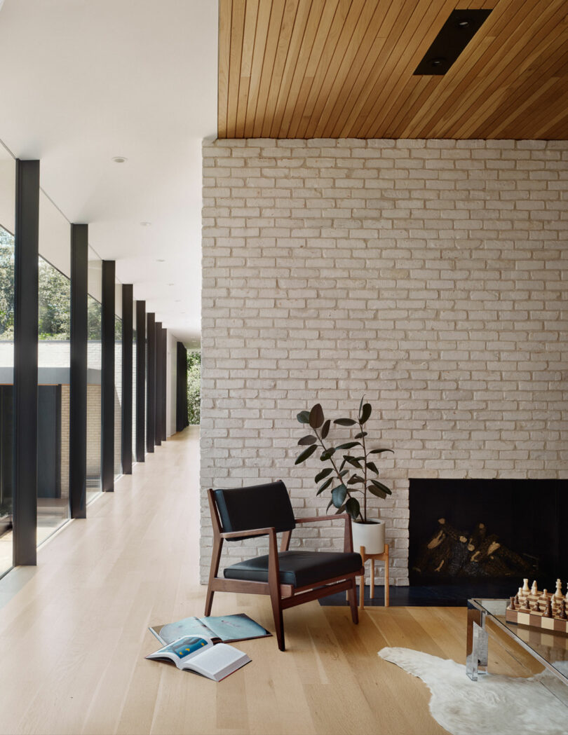 Modern room with a brick wall and fireplace, wooden ceiling, floor-to-ceiling windows, a black chair, plant, open books on the floor, and a chessboard on a glass table.