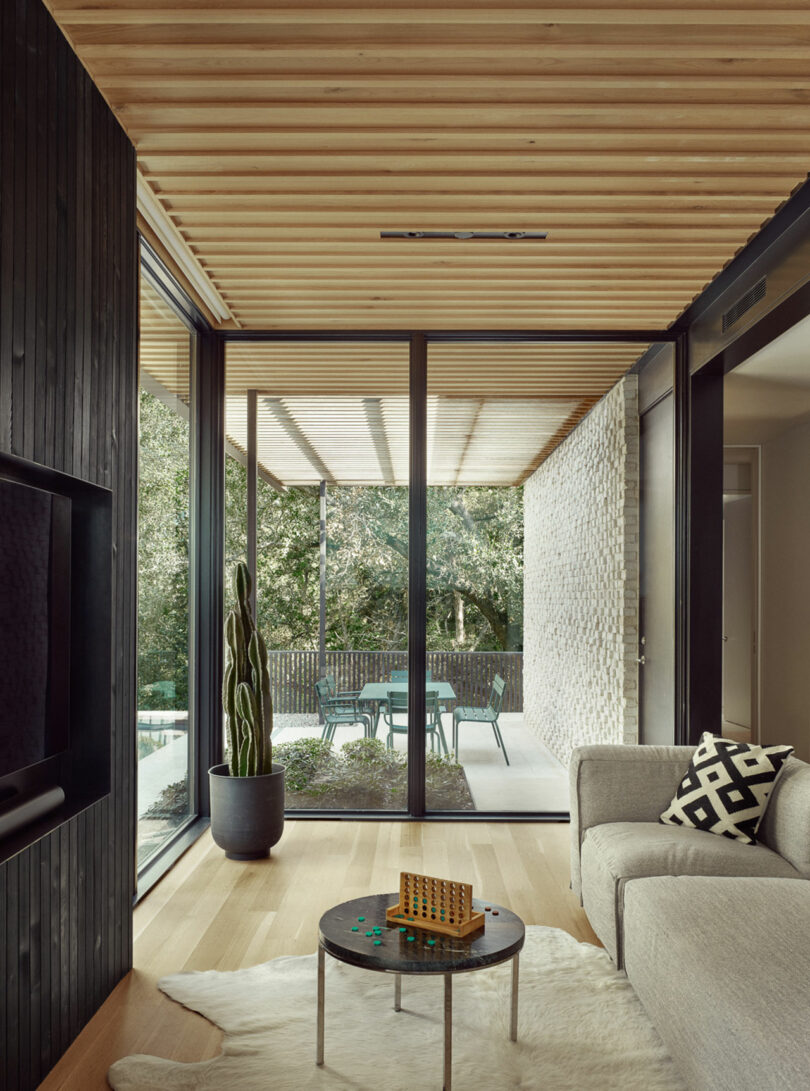 Sala de estar moderna con grandes puertas de vidrio, techo de madera y pisos de color claro. Un sofá gris con una almohada geométrica está al lado de una mesa redonda con un pequeño juego. Un patio es visible afuera.