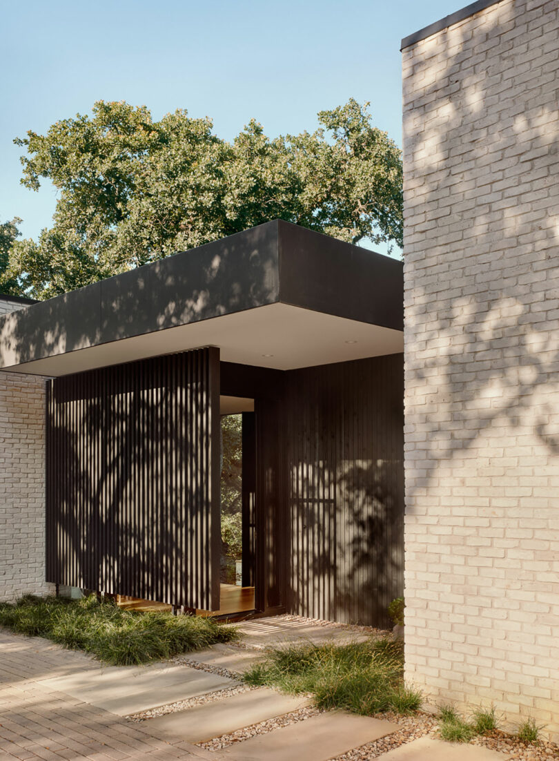 Entrada moderna de la casa con listones de madera verticales, paredes de ladrillo blanco y un techo plano. Sombras de árboles visibles en las paredes.