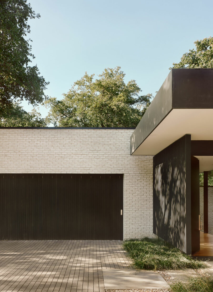 El exterior moderno de la casa con ladrillos blancos y acentos negros, con una gran puerta de garaje, techo plano y camino de entrada pavimentado enmarcado por hierba, debajo del cielo azul claro y rodeado de árboles.