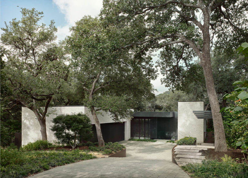 Casa moderna rodeada de árboles, con paredes de ladrillo blanco y grandes ventanas de vidrio, con un camino de entrada pavimentado que conduce a la entrada.