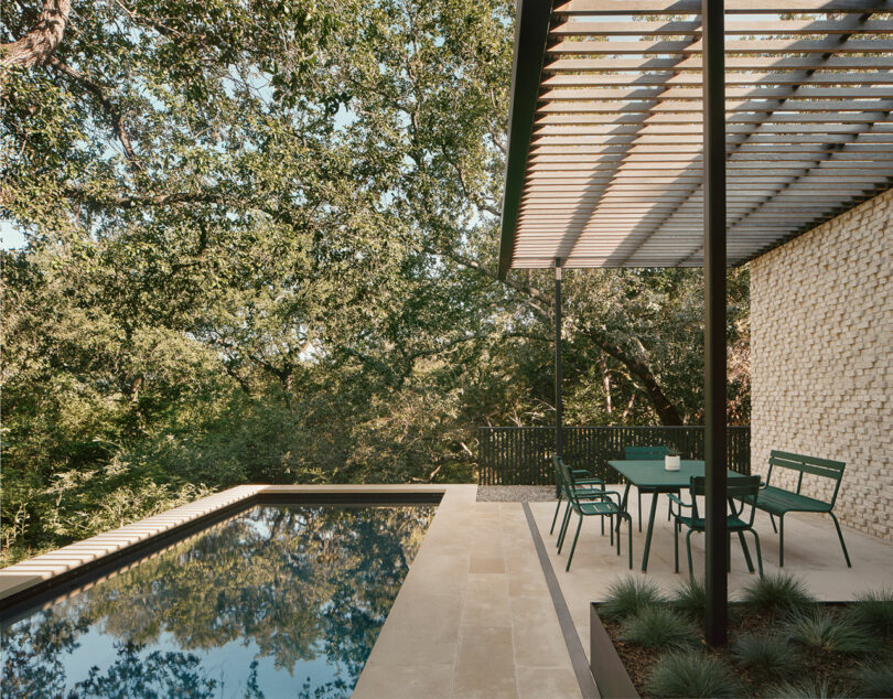 Un patio moderno al aire libre cuenta con una piscina rectangular, mesa y sillas verdes, y una cima de la pérgola, rodeada de exuberantes árboles.