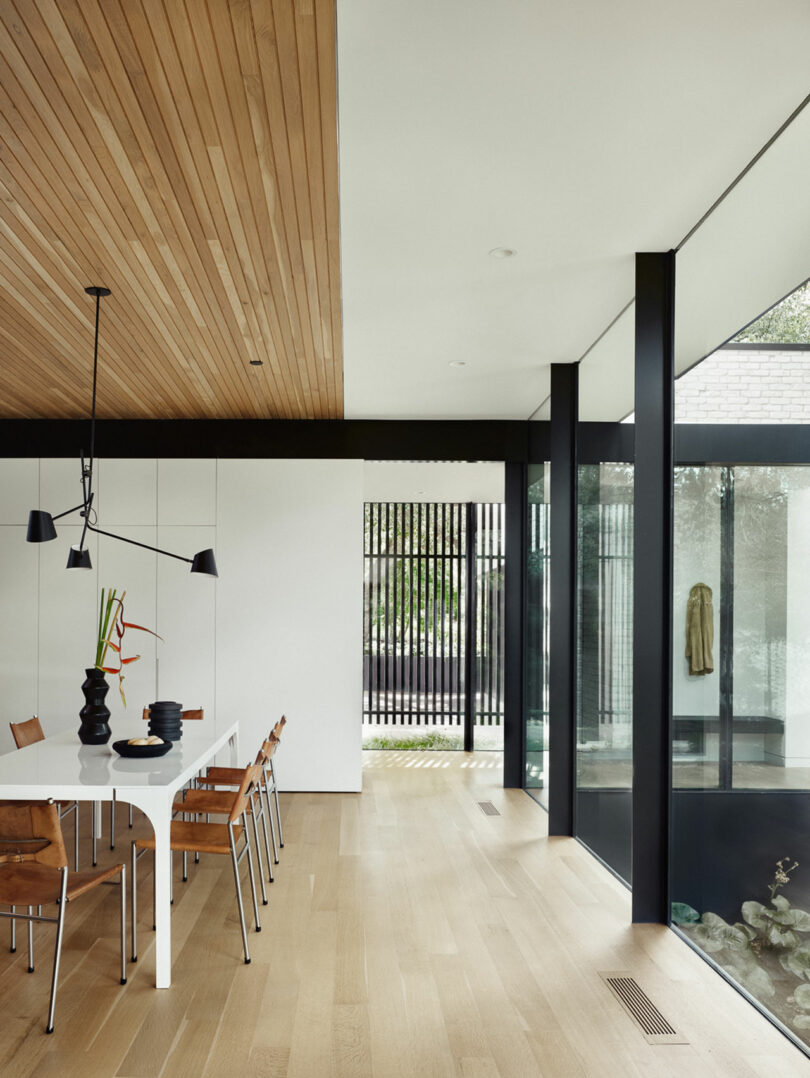 Moderno comedor con una mesa blanca, sillas de madera, luz colgante negra y un techo con panel de madera. Grandes ventanas y una pantalla decorativa de listones ofrecen vistas de la vegetación afuera.