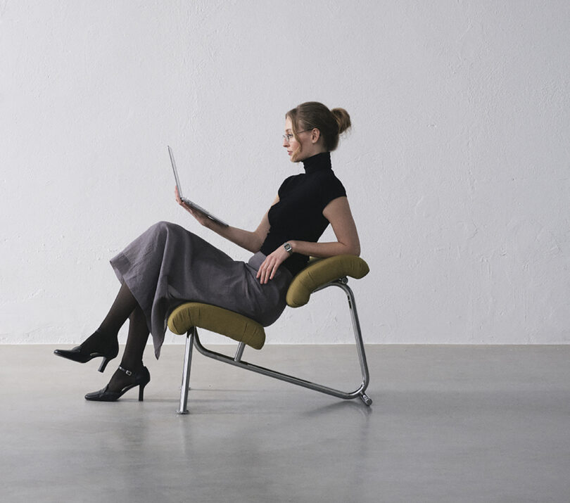 Woman in a modern chair, wearing a black top and gray skirt, looks at a laptop in a minimalist room.