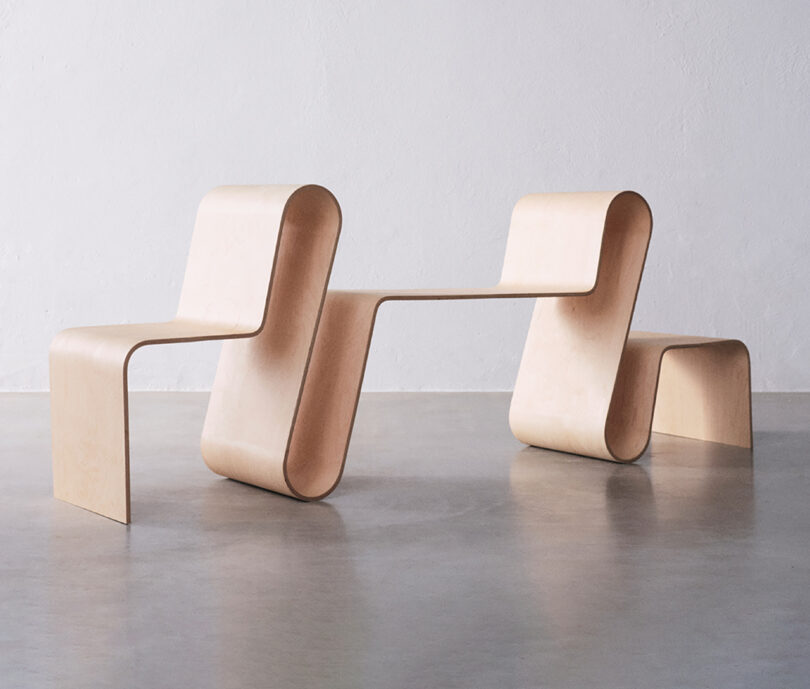 Sculptural wooden benches with a flowing, wave-like design set against a plain white wall and gray floor.