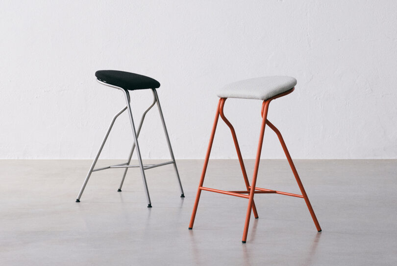 Two minimalist stools with sleek metal frames and simple seats stand on a smooth, light-colored floor against a plain white wall.