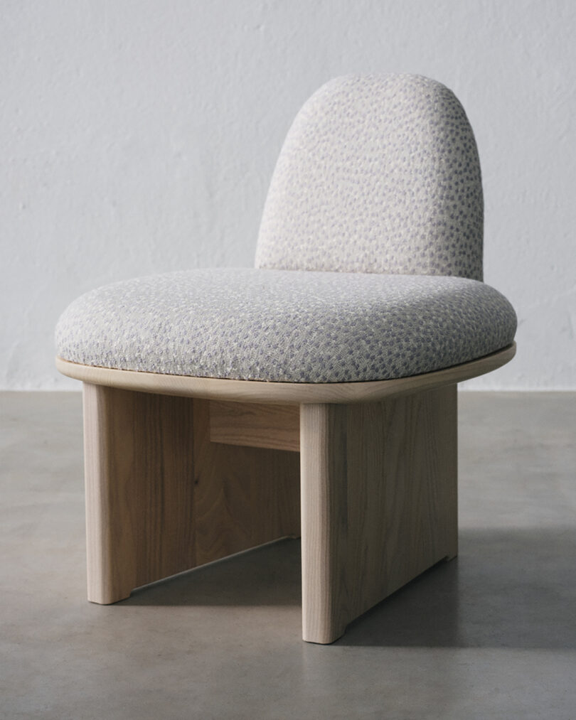 Simple wooden chair with a rounded backrest and cushioned seat upholstered in light gray fabric, placed on a light-colored floor against a plain wall.