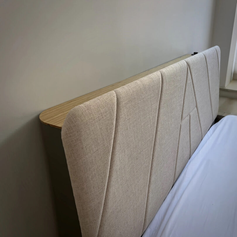 Beige upholstered headboard with vertical stitching, attached to a bed.