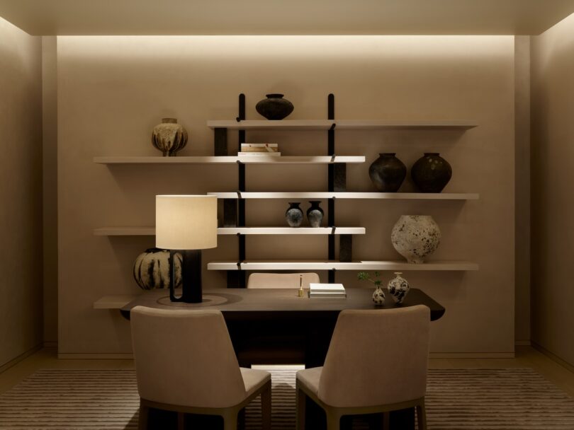 A dimly lit room features a table with two chairs, a lit lamp, and shelves with pottery.