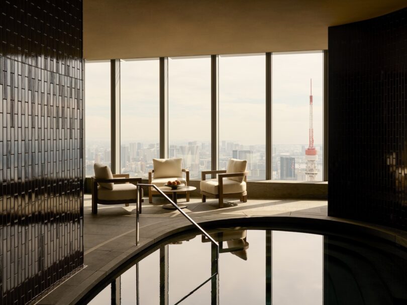 Indoor pool area with two chairs and a table facing large windows. Cityscape and a red-and-white tower are visible in the background.