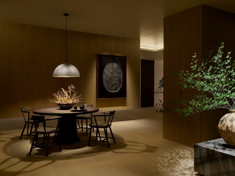 A modern dining room with a round table, five chairs, large pendant light, decorative branches in the center, a textured artwork on the wall, and a vase with greenery on the side.