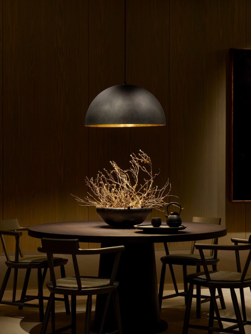 A round table with chairs is illuminated by a pendant lamp. A bowl with dry branches serves as a centerpiece, and a teapot with a cup is placed on the table.