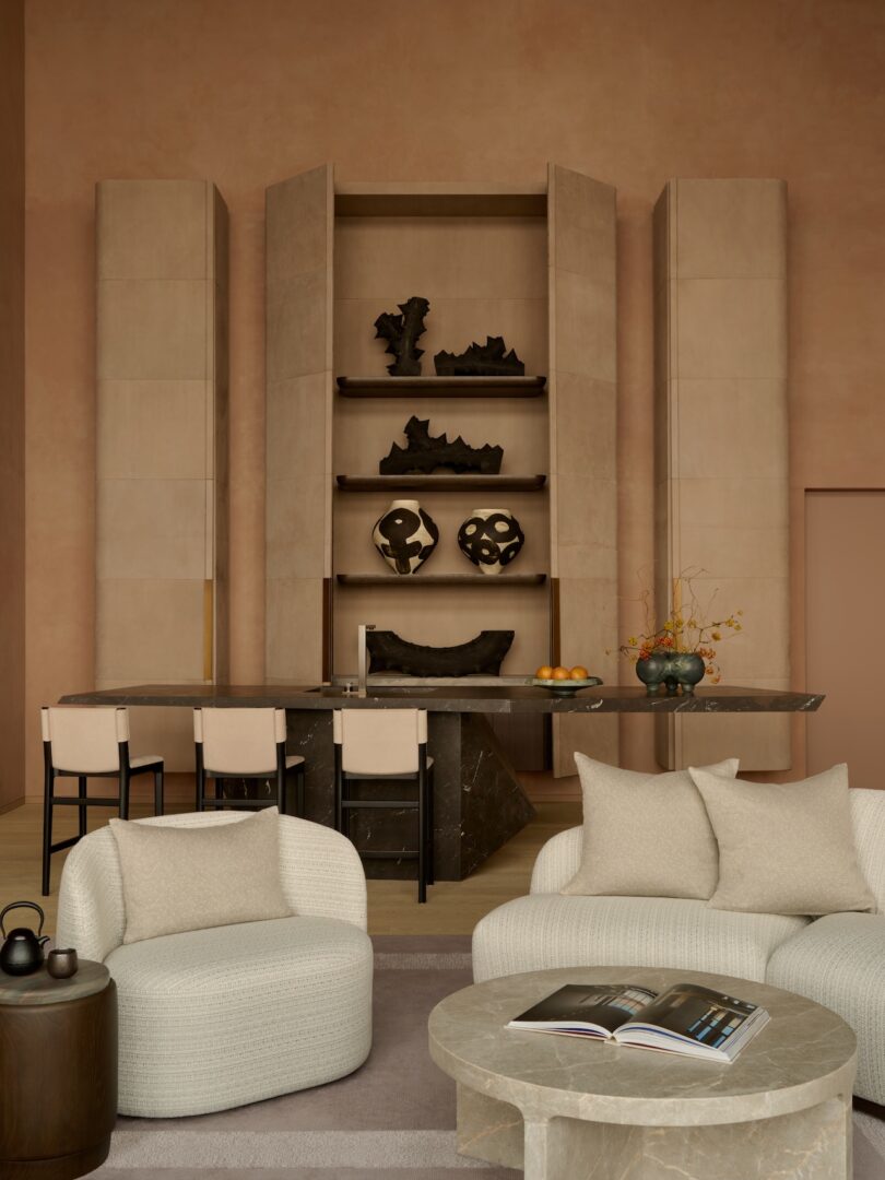 Modern living room with beige chairs, round marble table, and open book. Shelves with abstract sculptures, a dark table, and four high chairs are in the background. Minimalist decor with warm tones.