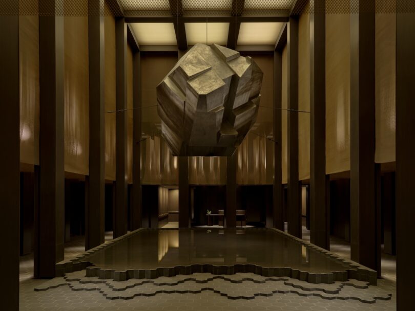 A large, geometric stone sculpture is suspended in a dimly lit room with tall columns and a pool reflecting its shape on the floor.