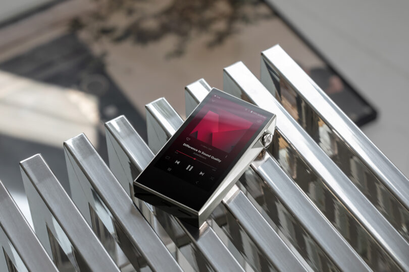 The SP3000M digital music player, with its sleek touchscreen display showing a song, rests elegantly on a shiny metal surface adorned with linear grooves.