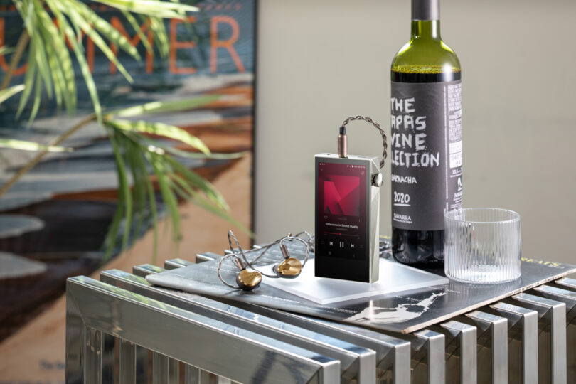 A digital device, model SP3000M, with earphones sits on a tray beside a wine bottle and an empty glass. Sunglasses rest next to the sleek gadget on the metallic surface.