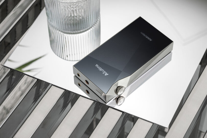 A sleek, silver SP3000M audio player with control knobs rests on a reflective surface beside a ridged glass of water.