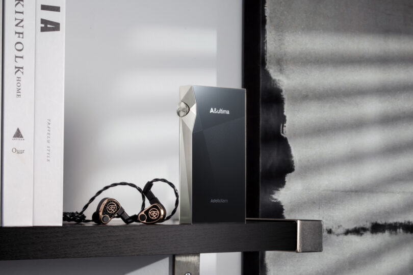 A sleek audio device, SP3000M, labeled "A&ultima" rests elegantly on a shelf beside books and a pair of stylish earphones.