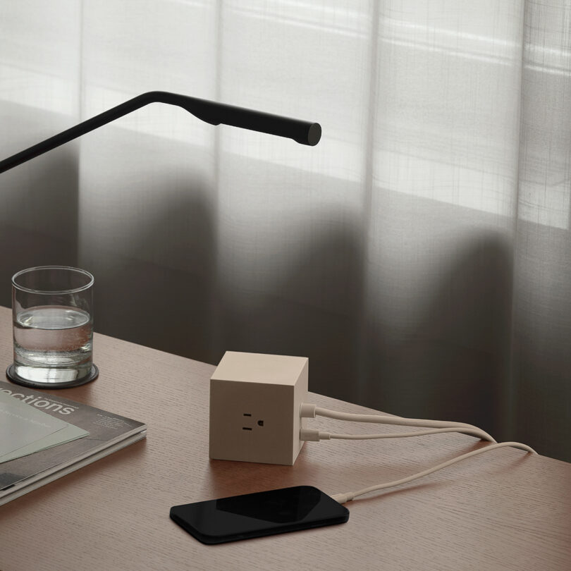 A desk features a modern lamp, a glass of water, an AVOLT Square 1 power adapter with two cords, a smartphone, and a closed magazine, with soft light filtering through a curtain in the background.