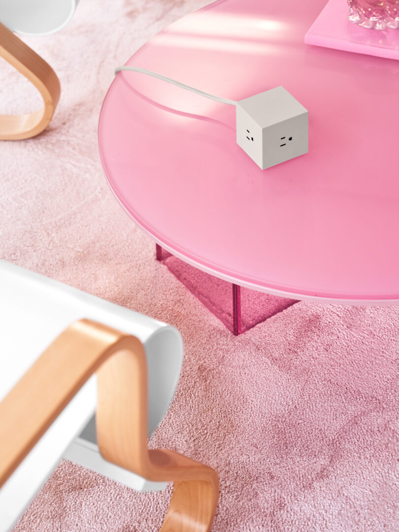 A pink round coffee table on a matching carpet, accompanied by a white AVOLT Square 1 power adapter and a rolled-edge chair with wooden armrests nearby.