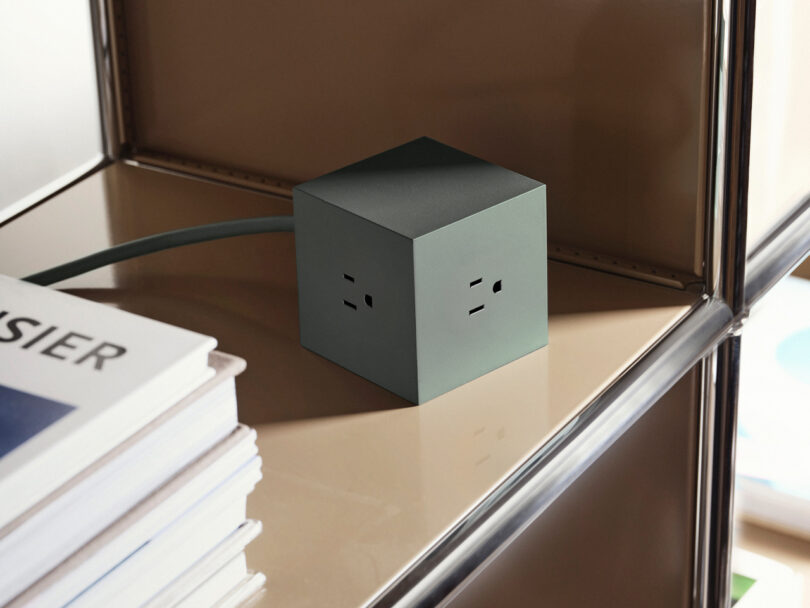 An AVOLT Square 1, a green cube-shaped power outlet, sits on a brown shelf next to a stack of books.