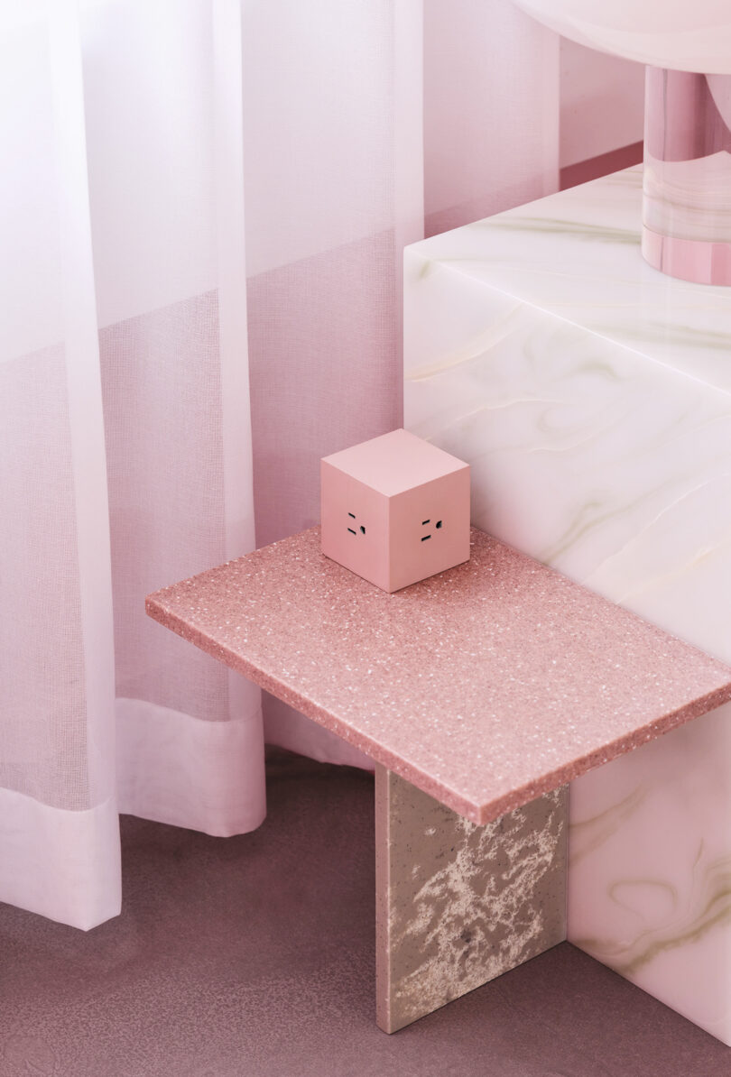 A pink cube-shaped AVOLT Square 1 sits on a pink terrazzo shelf beside a white and pink veined marble surface. A sheer curtain is partially visible in the background, adding an element of subtle elegance to the scene.