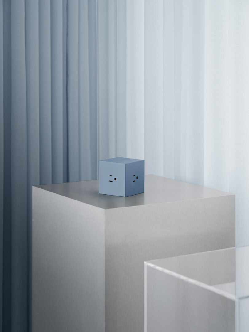 The AVOLT Square 1, a blue cube with face-like features, sits elegantly on a gray pedestal against light blue curtains.