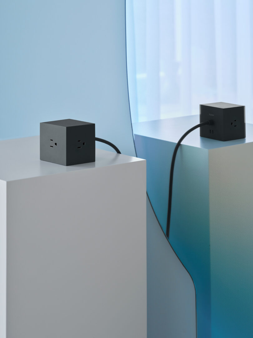 The AVOLT Square 1, a sleek black cube-shaped power outlet, rests elegantly on a white pedestal. Its multiple sockets and stretching power cord are artfully reflected in the curved mirror beside it.