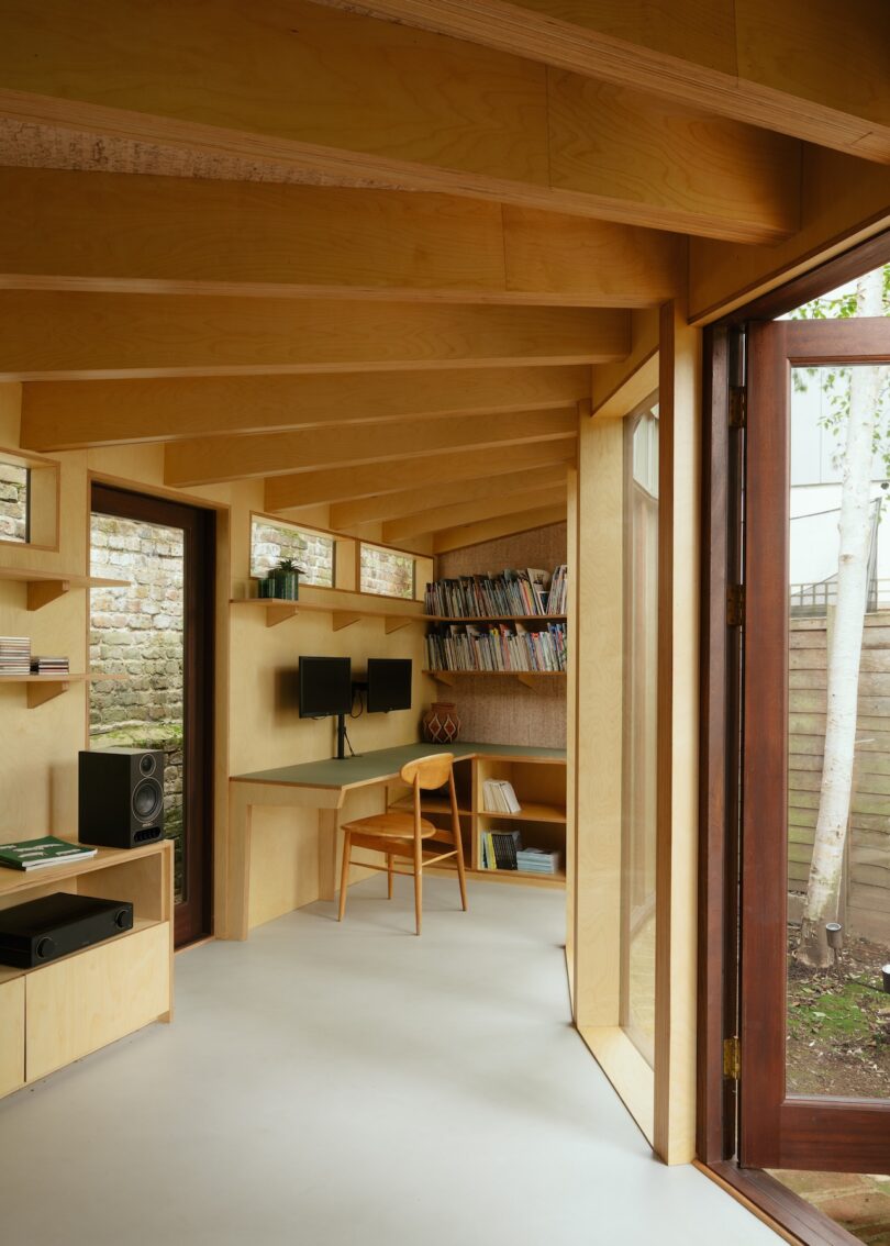 A modern wooden interior office with a desk, chair, dual monitors, and bookshelves. Large door open to a fenced yard with birch trees.