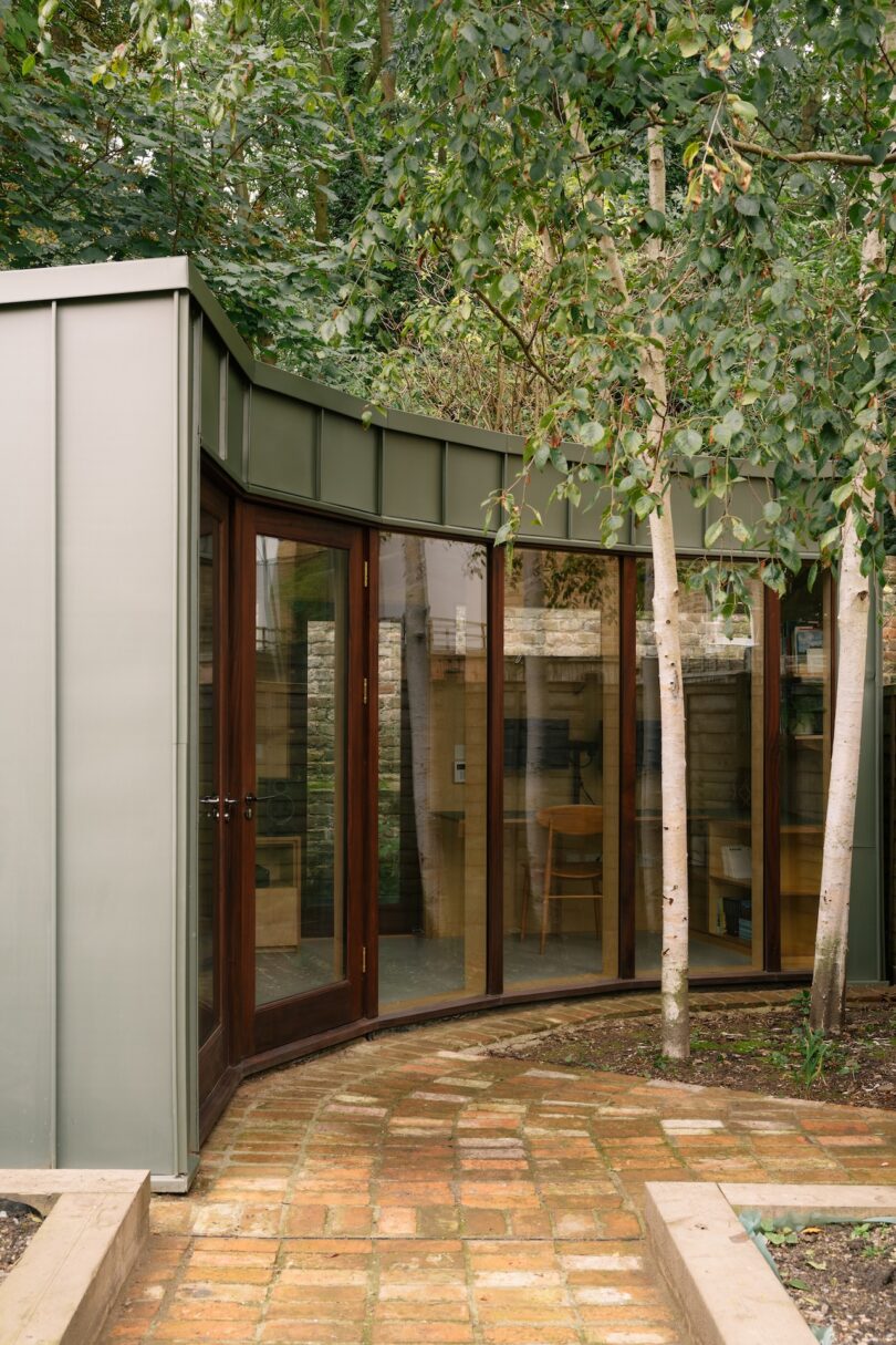 Modern glass-walled garden room with wooden doors, surrounded by trees and situated on a brick path, under a metal roof.