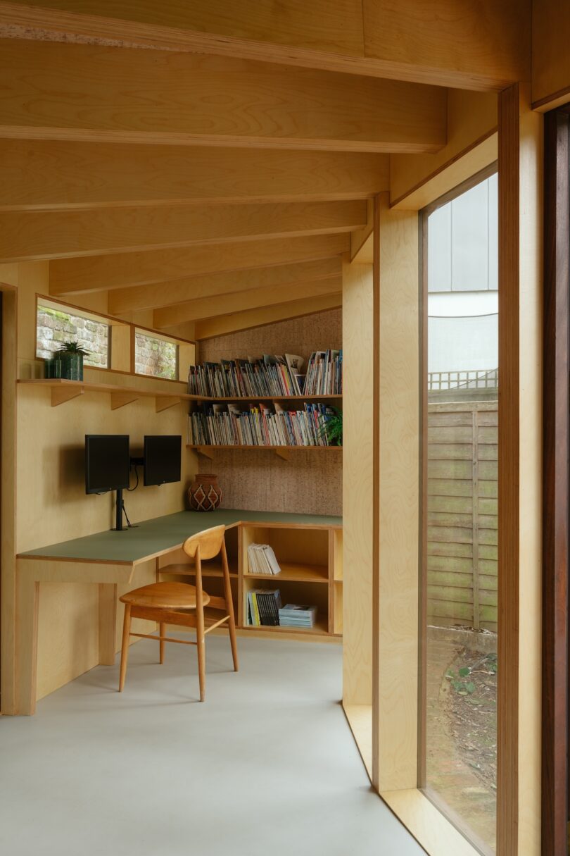 Wooden home office with large window, chair, desk with dual monitors, bookshelves filled with books, and a potted plant.